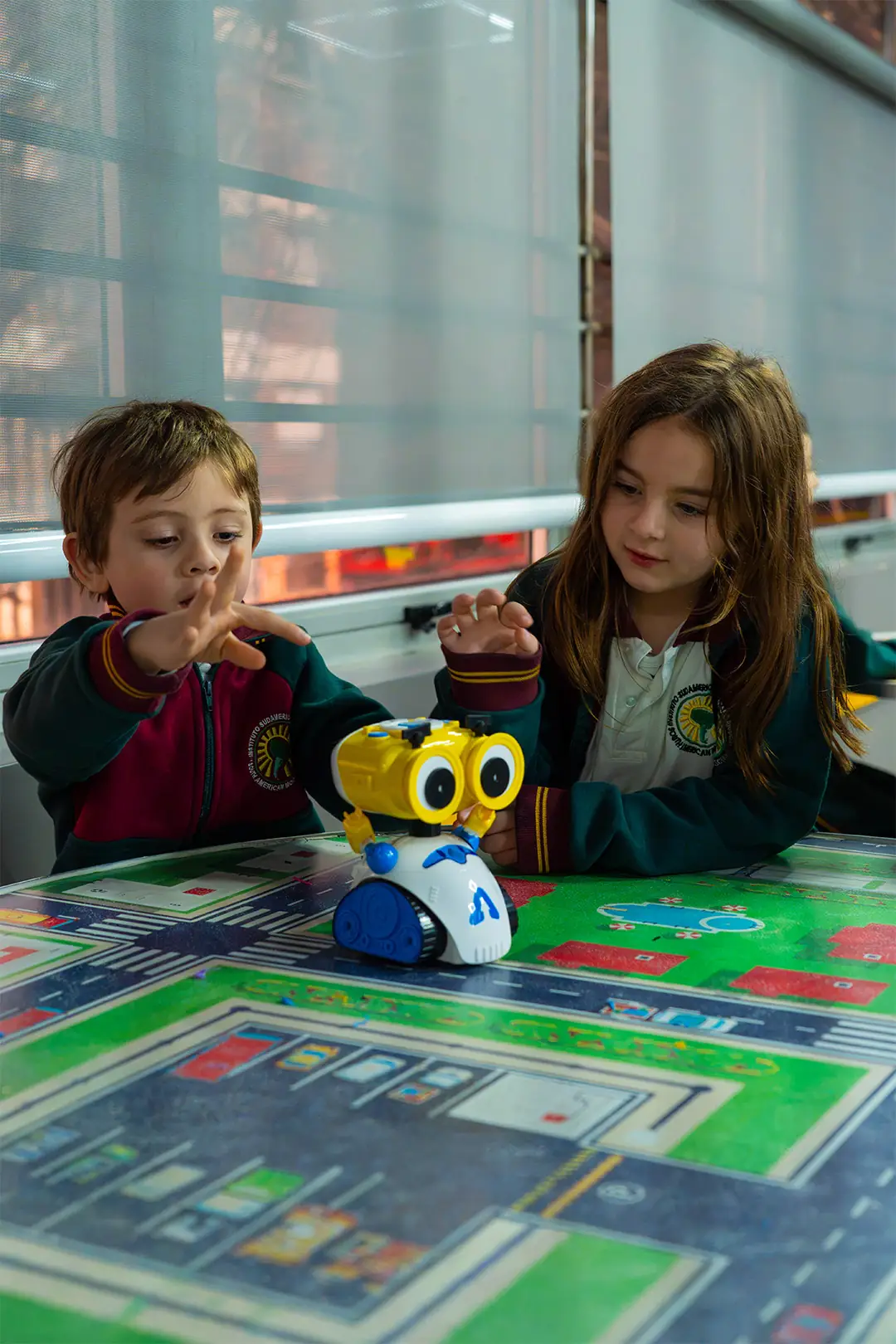 Dos niños del Jardín de Infantes del IESM sentados a la mesa con un tablero de actividades y un robot de piso
