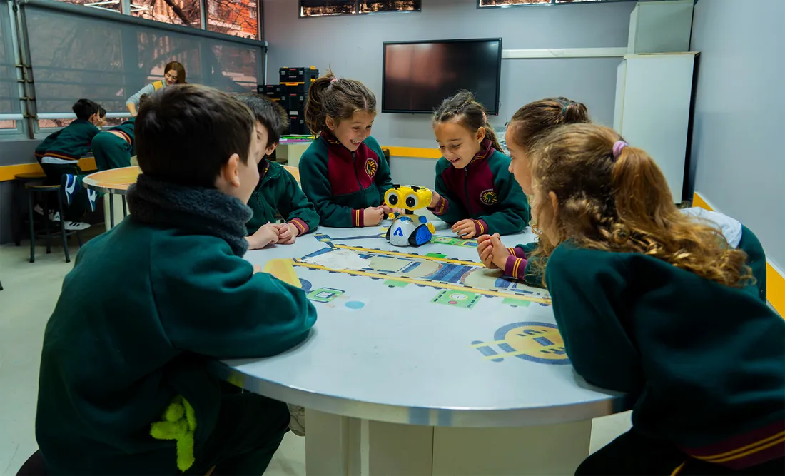 Alumnos del Nivel Primario del IESM trabajando en grupo alrededor de una mesa con un tablero de actividades y un robot de piso